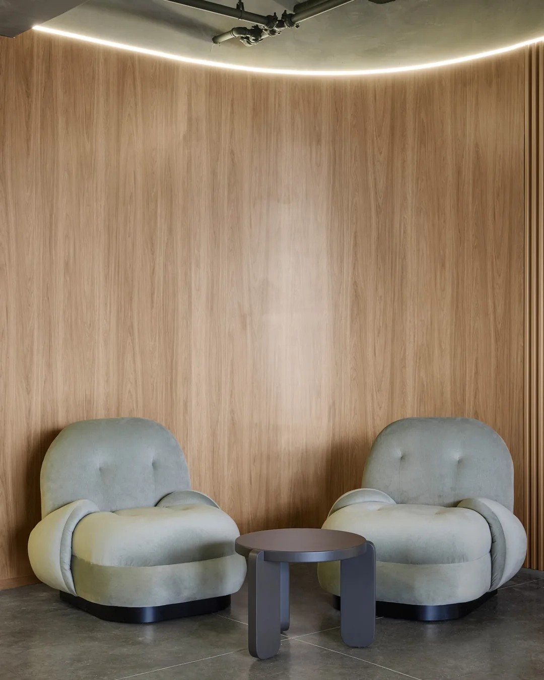 Sillones tapizados en verde con mesa auxiliar circular negra frente a panel en madera de diseño cont