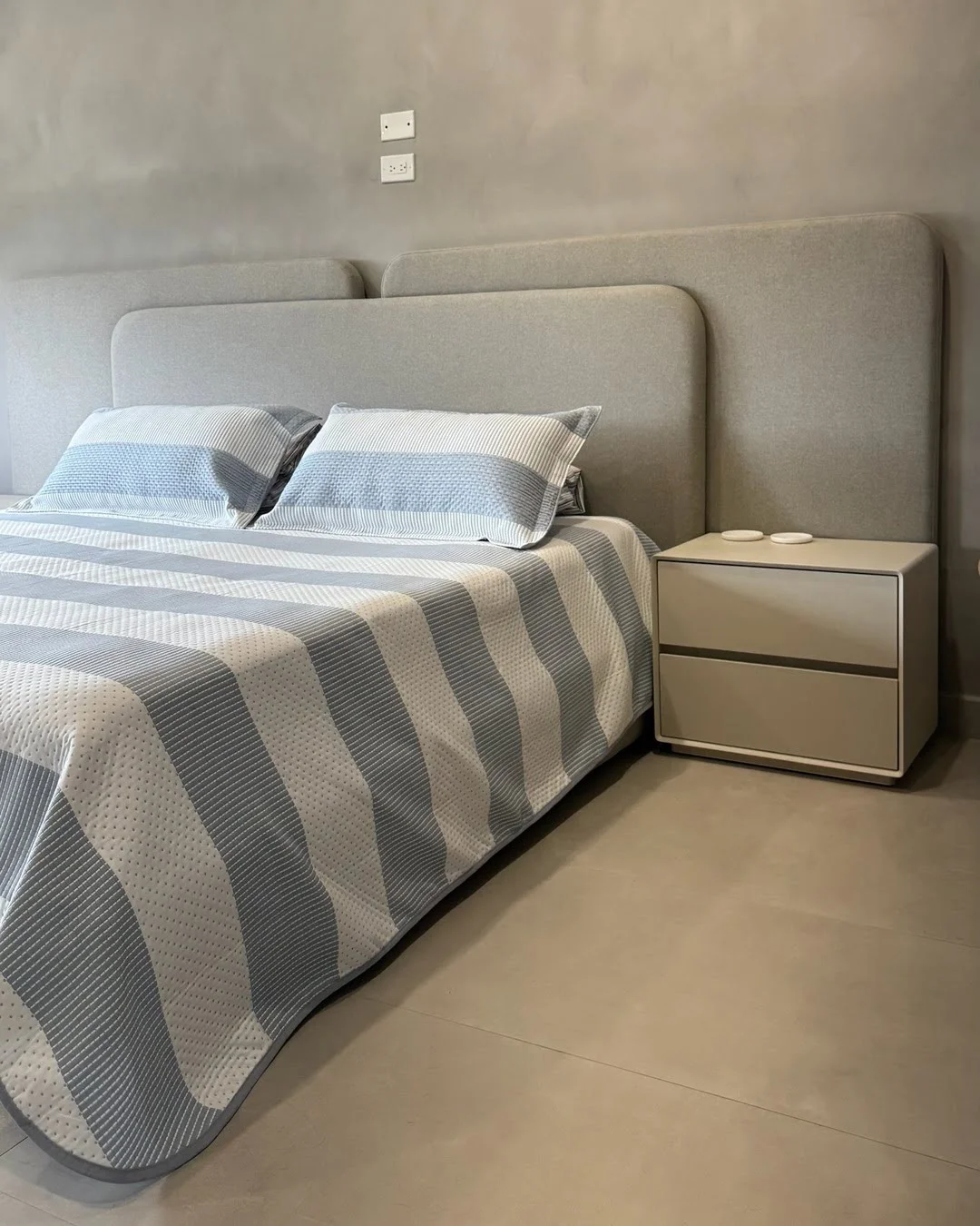 Habitación principal con cama de cabecero acolchado gris y mesa de noche minimalista.